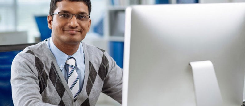 A student taking Online Career Training Programs sitting in front of his computer. He is smiling and looking at the camera.